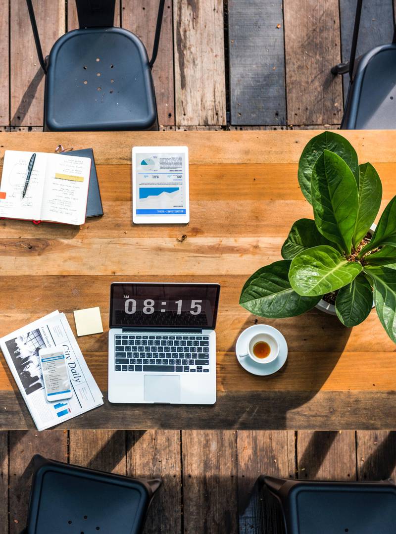 Overhead view of a wooden desk with a laptop, tablet, notebook, coffee and a potted plant